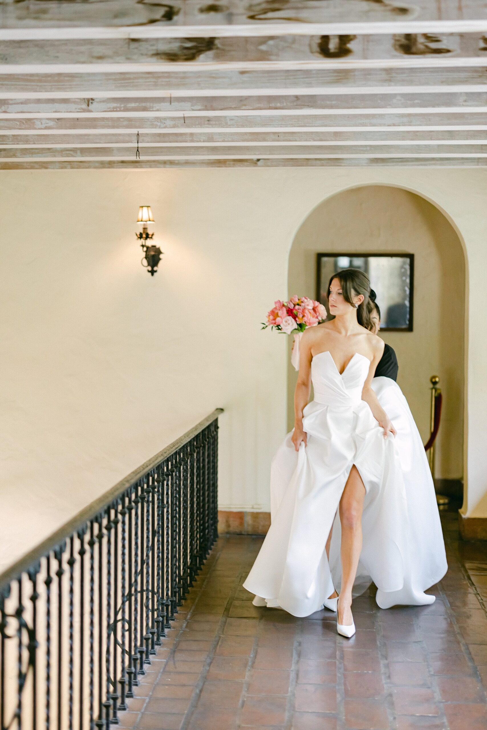 Bride walking towards her first look with the groom assisted by makeup artist holing her dress and blush bouquet