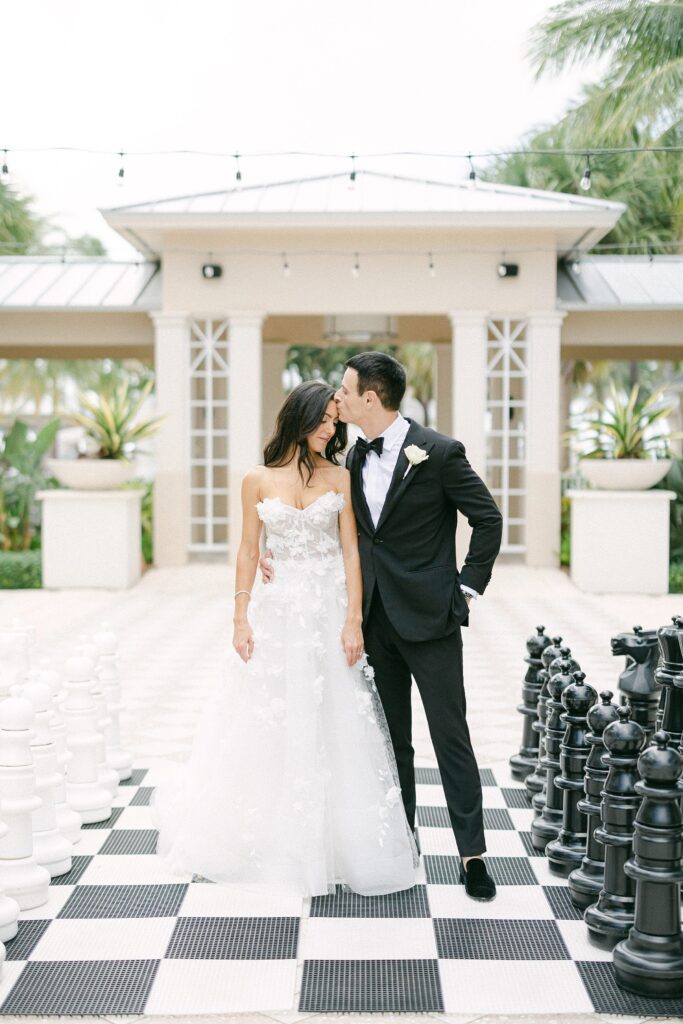 bride and groom standing in the middle of a giant chess board while kissing her forehead