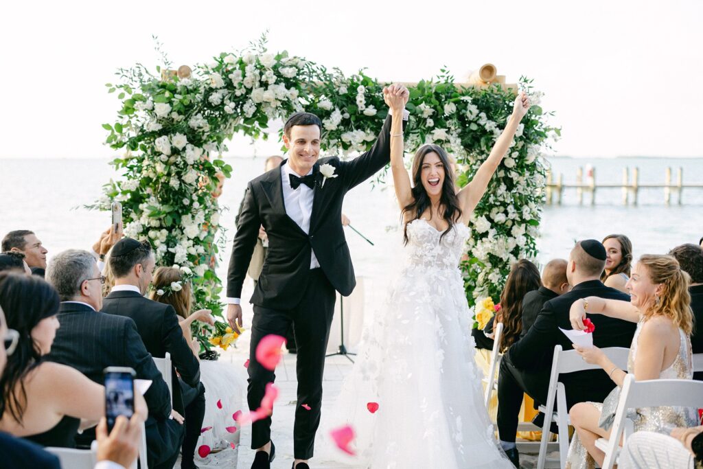 Bride and groom hold hands in the air as guests throw pink petals down the aisle