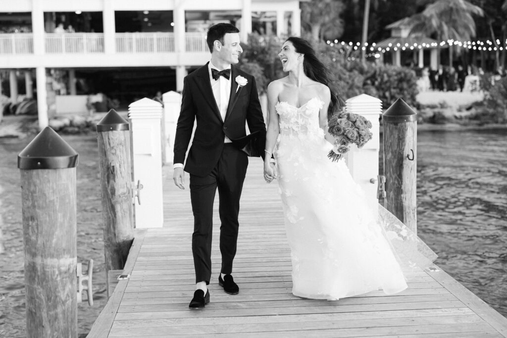 black and white photo of newlyweds laughing happily as they walk down the pier into the sunset