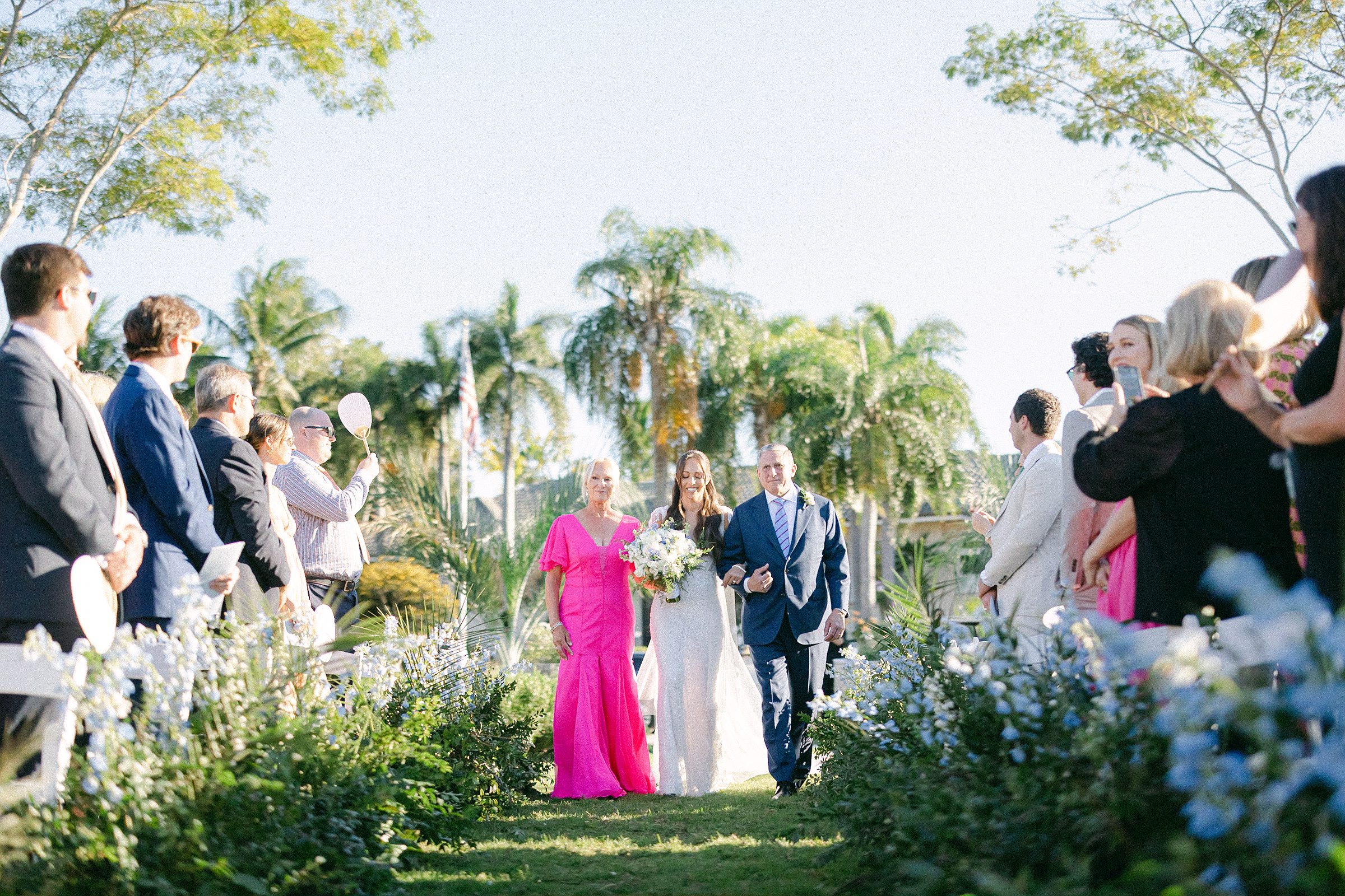 the bride walking down the aisle escorted by her father in a blue suite and mother in a pink dress