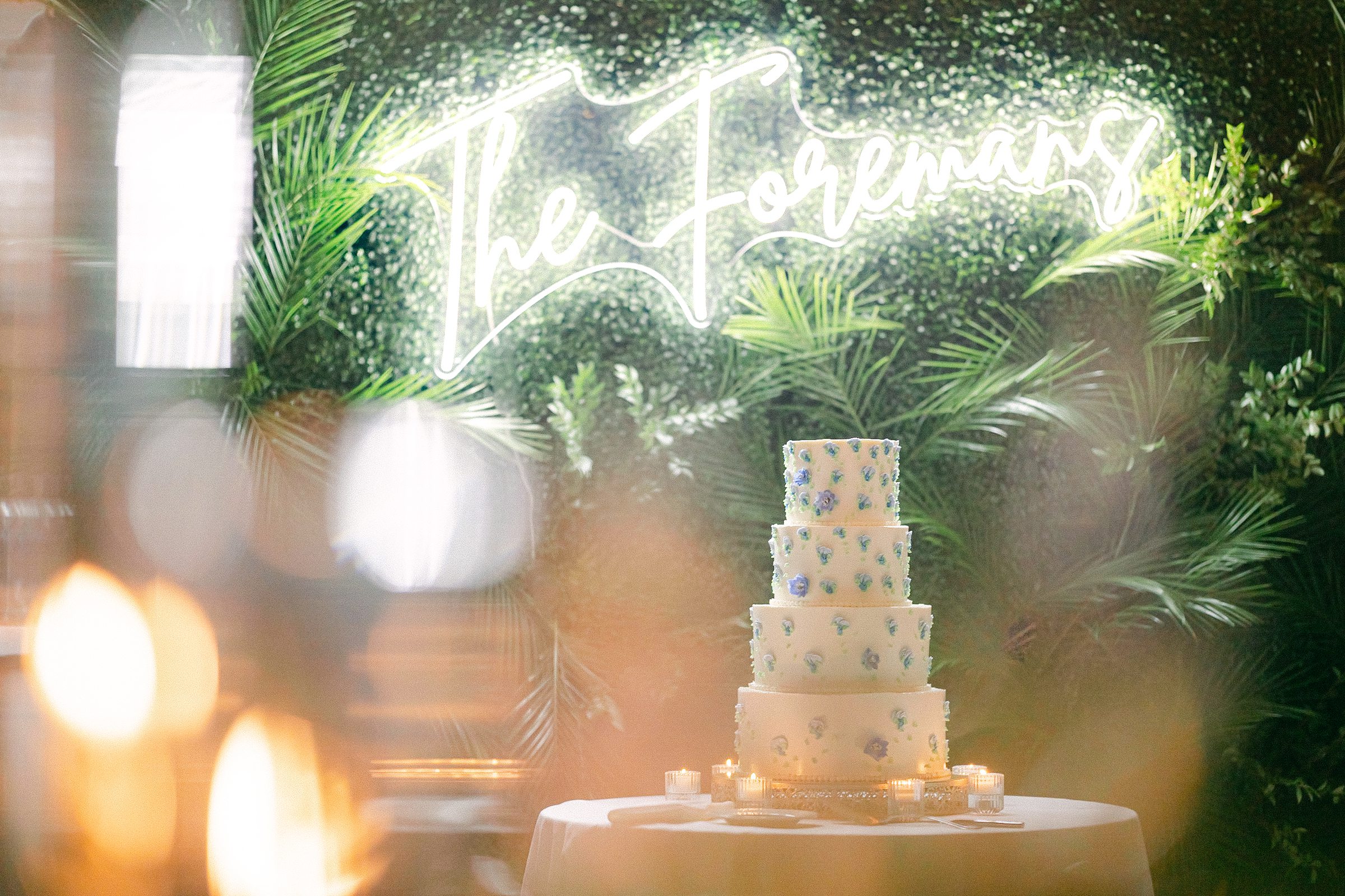 an artistic shot of the 4 tiered cake in front of a green wall with a neon sign above reading The Foremans