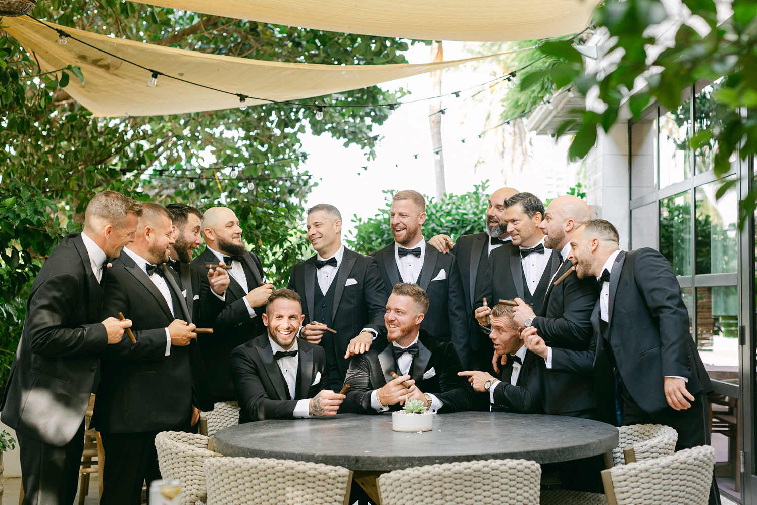 groom and groomsmen siting together ina group laughing while smoking cigars