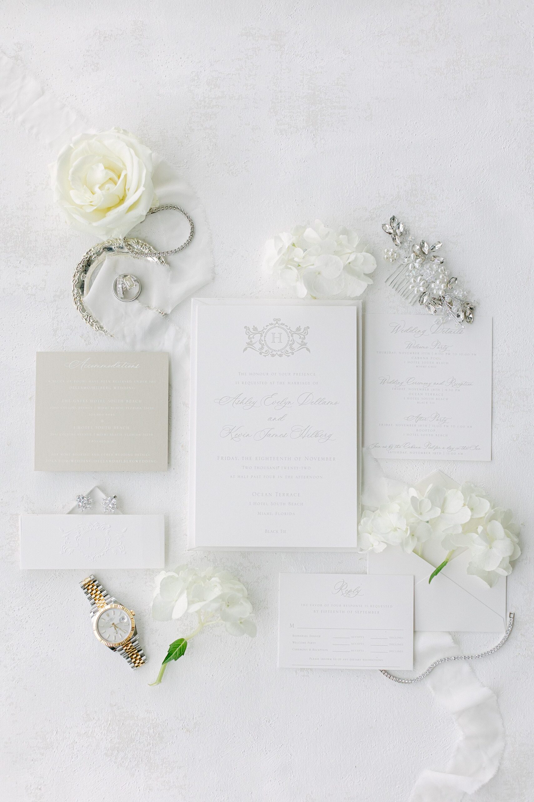 White Lay flat of bridal details including the wedding invitation, hair piece, wedding bands, diamond earrings and rolex watch