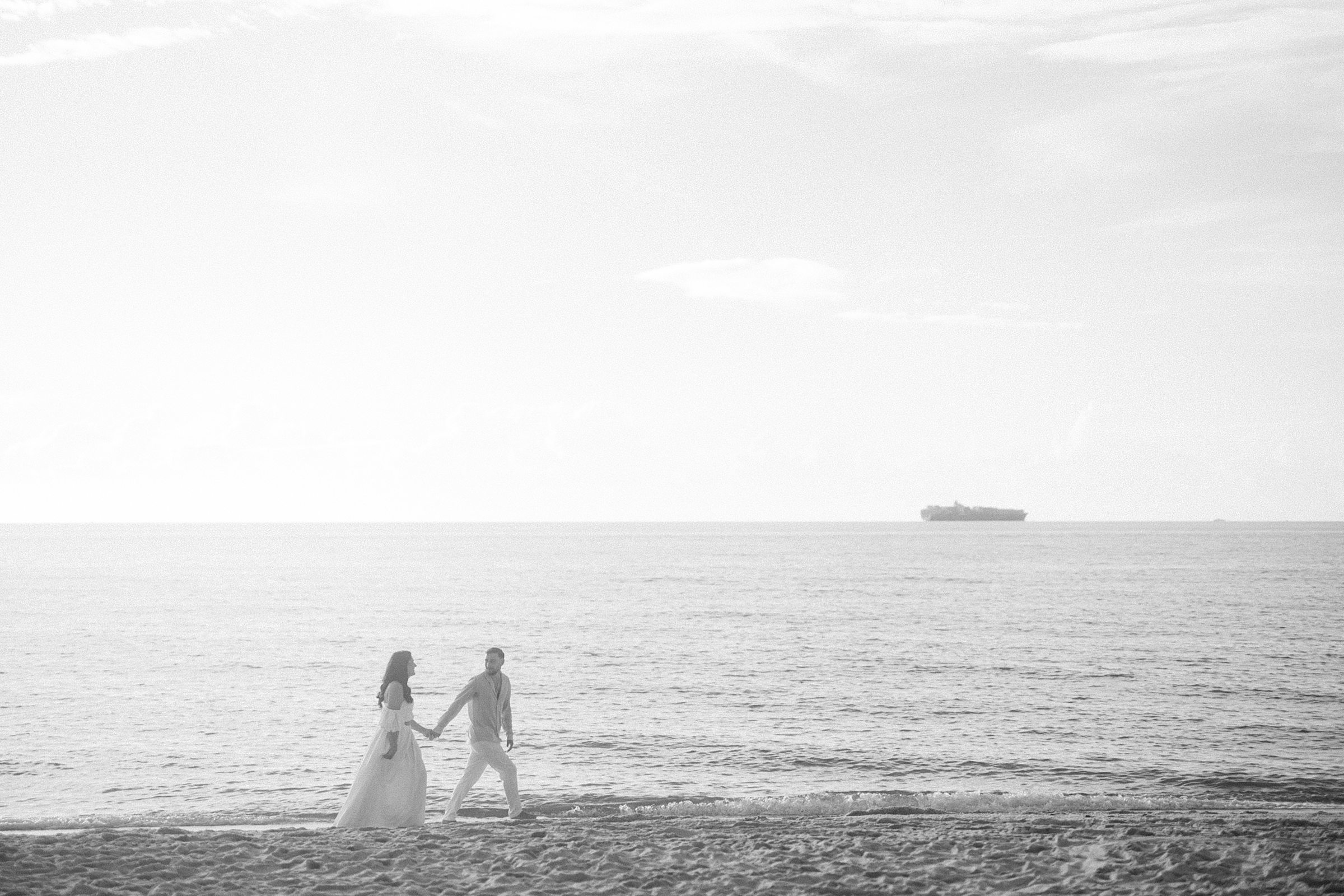 Black and white image of a couple walking down the beach with a cruise ship in the fair distance