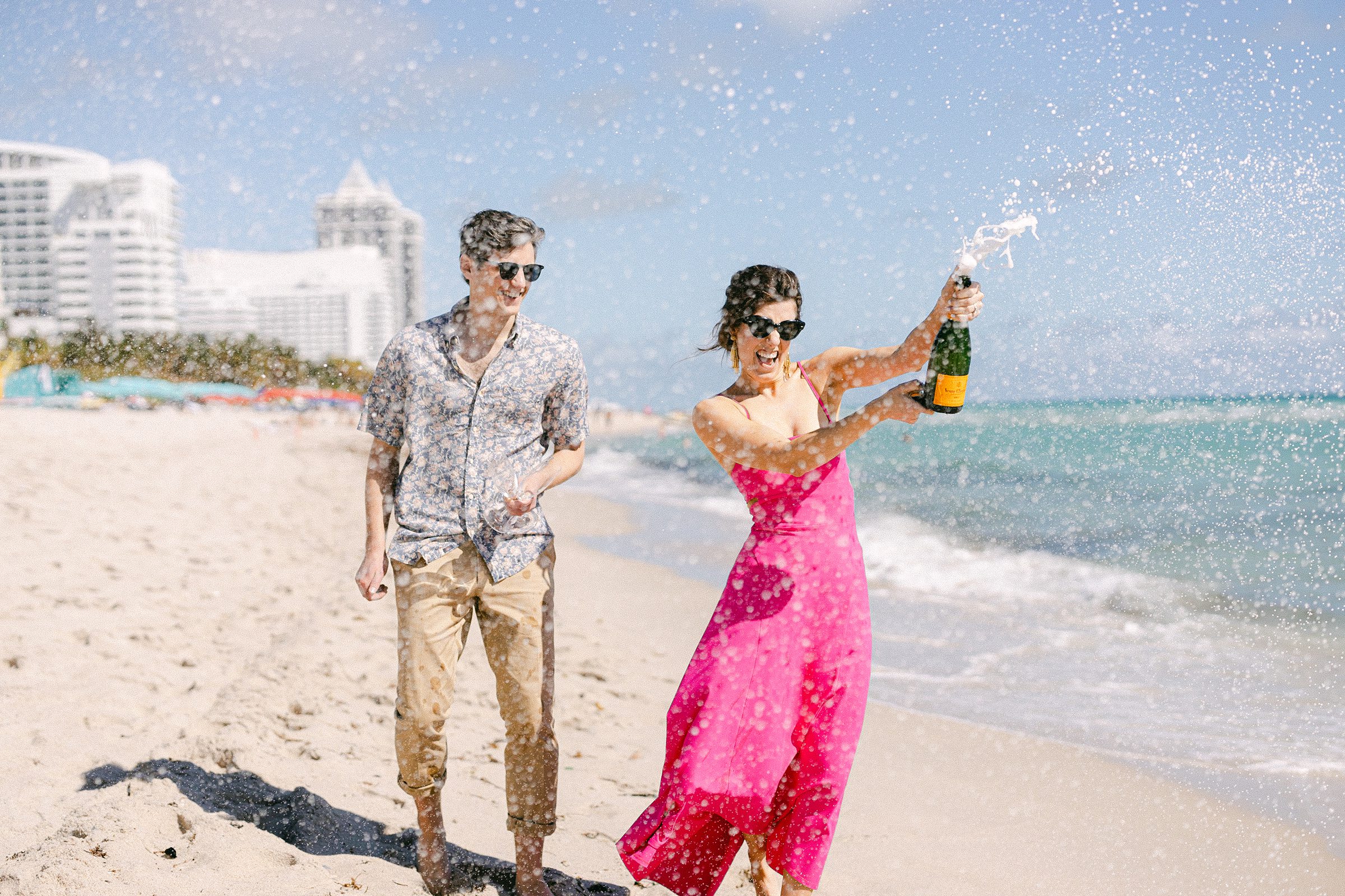 engaged couple spraying champagne at The Confidante Miami Beach. She's wearing a hot pink dress. He is wearing a floral shirt and khaki pants