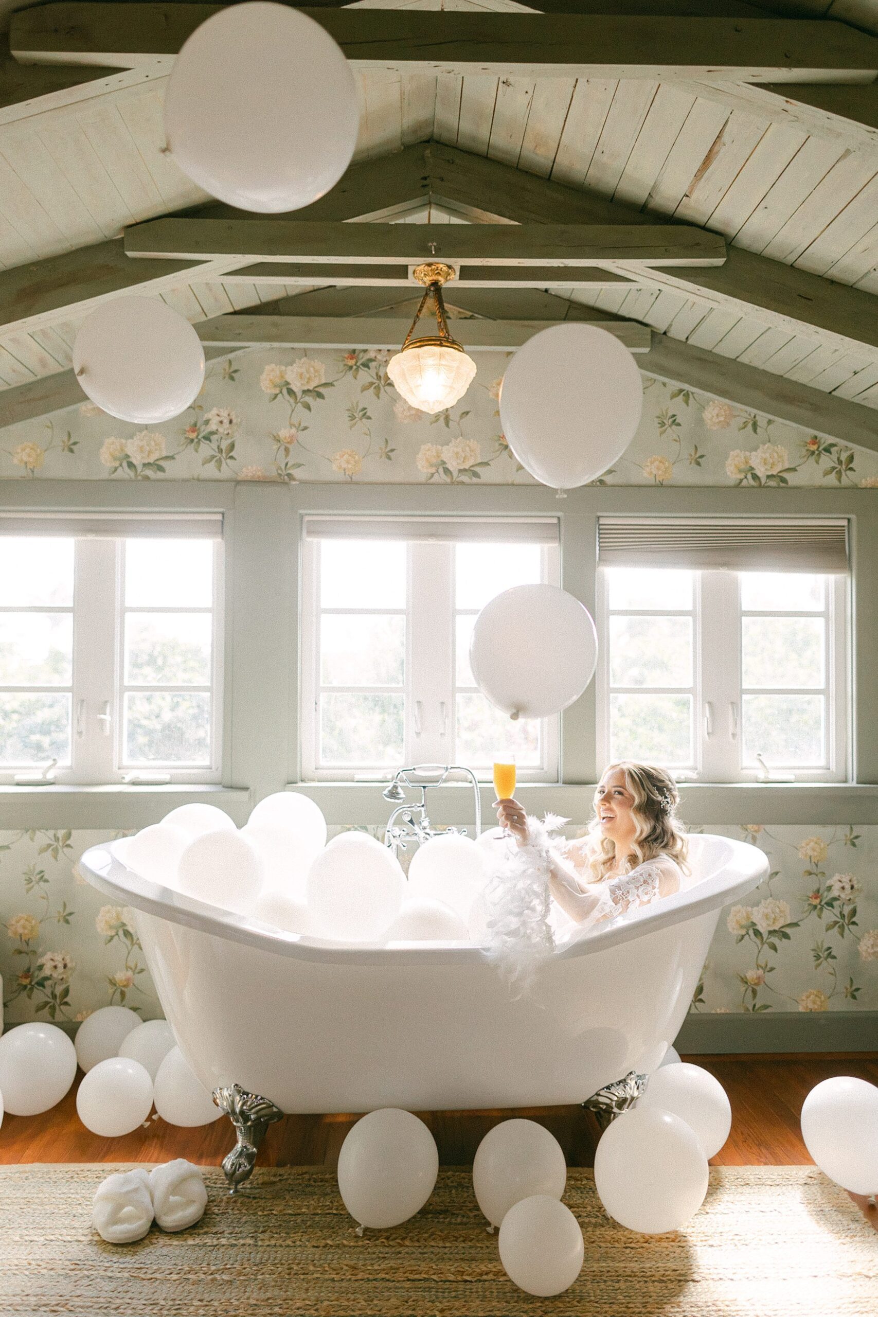 Bride in bath tub with an orange mimosa and white balloons in the air.